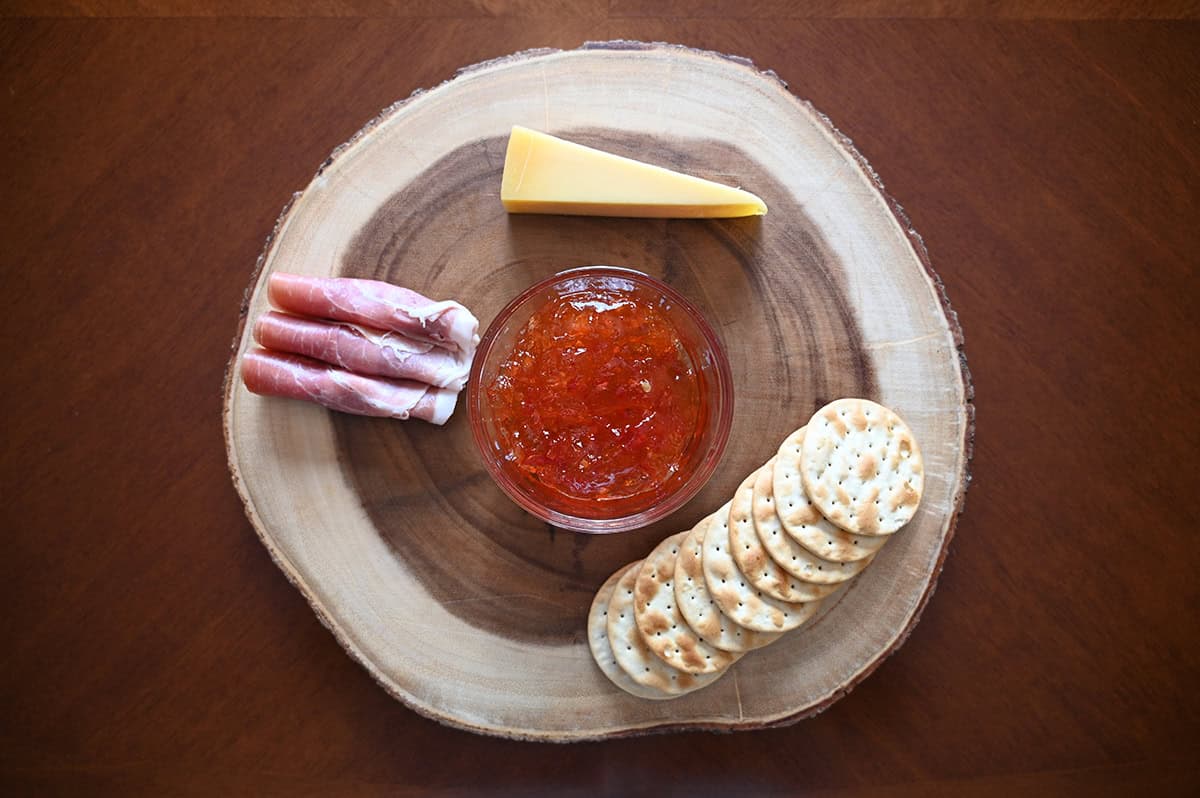 Image of a charcuterie board with cheese, prosciutto, crackers and red chili pepper spread on it.