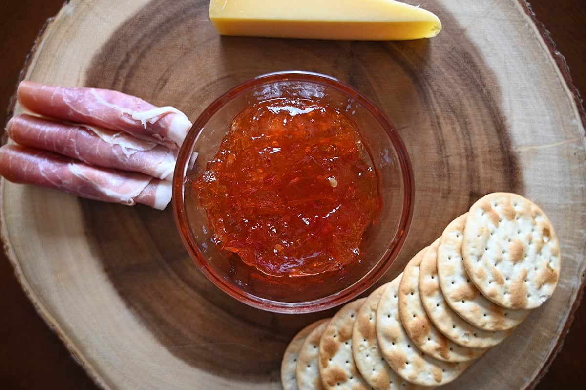 Closeup image of the red chili pepper spread in a small dish on a charcuterie board with crackers, meat and cheese on it. 