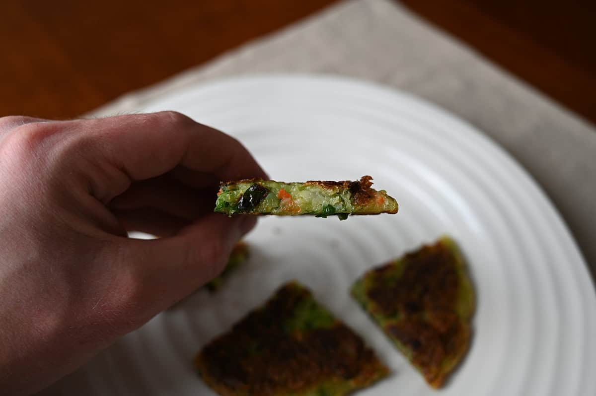 Closeup sideview image of a hand holding one piece of vegetable pancake so you can see how thick it is and what the center looks like.