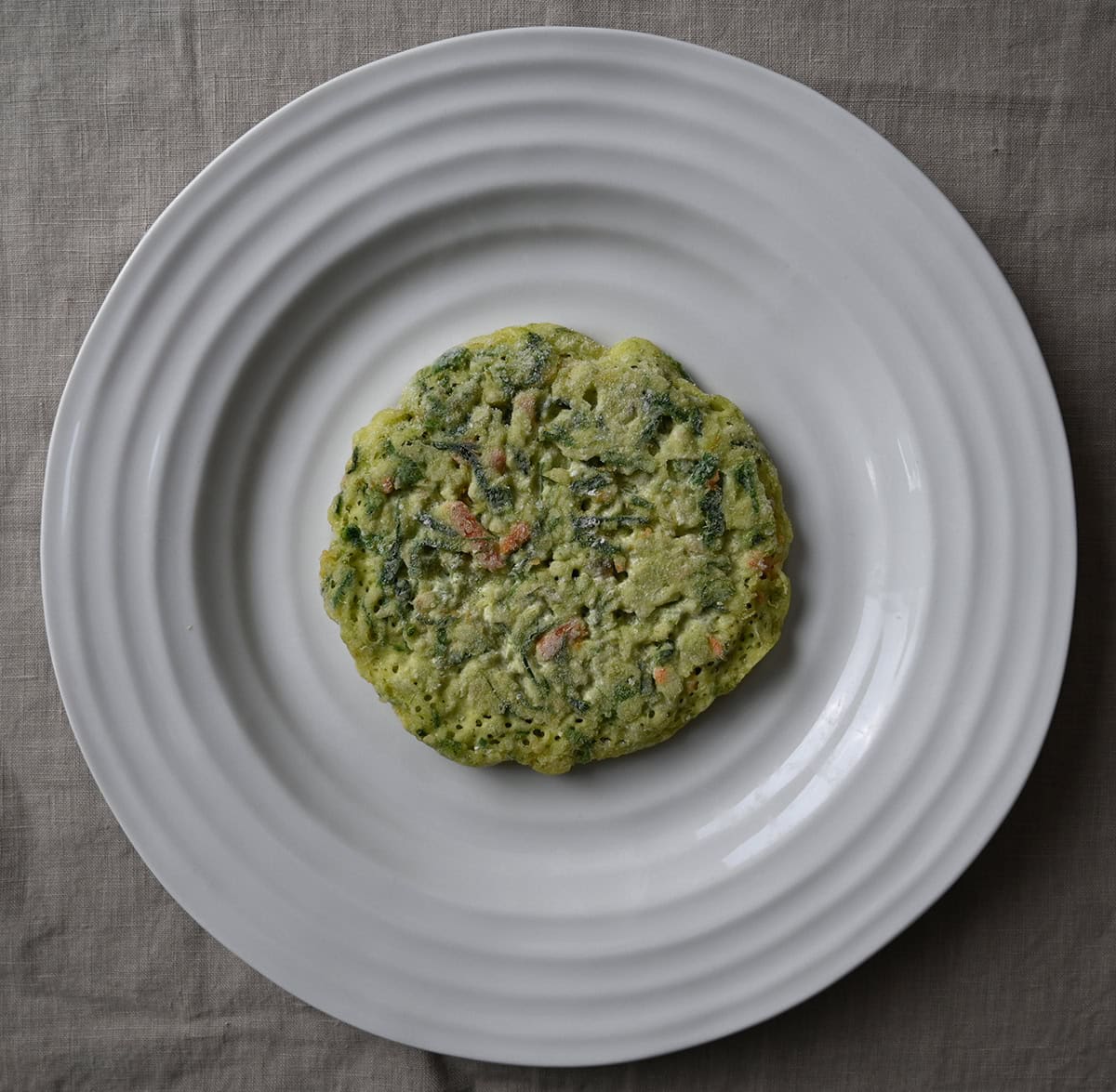Image of one frozen uncooked vegetable pancake sitting on a white plate. 