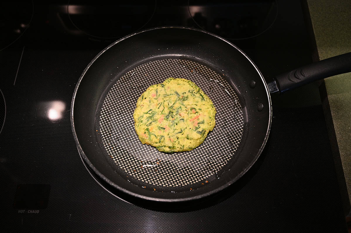 Image of one vegetable pancake being cooked in oil in a frying pan on the stovetop.