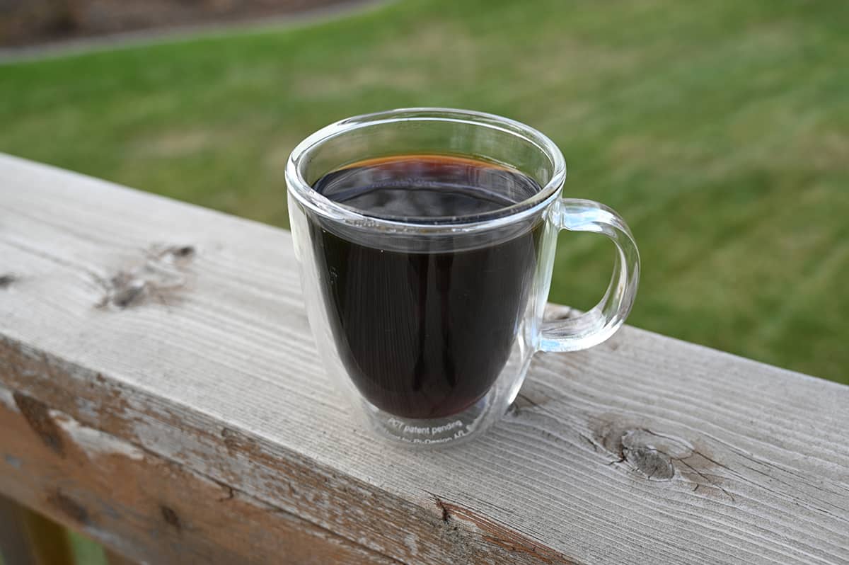 Image of a clear glass mug of black coffee sitting on a deck with grass in the background.