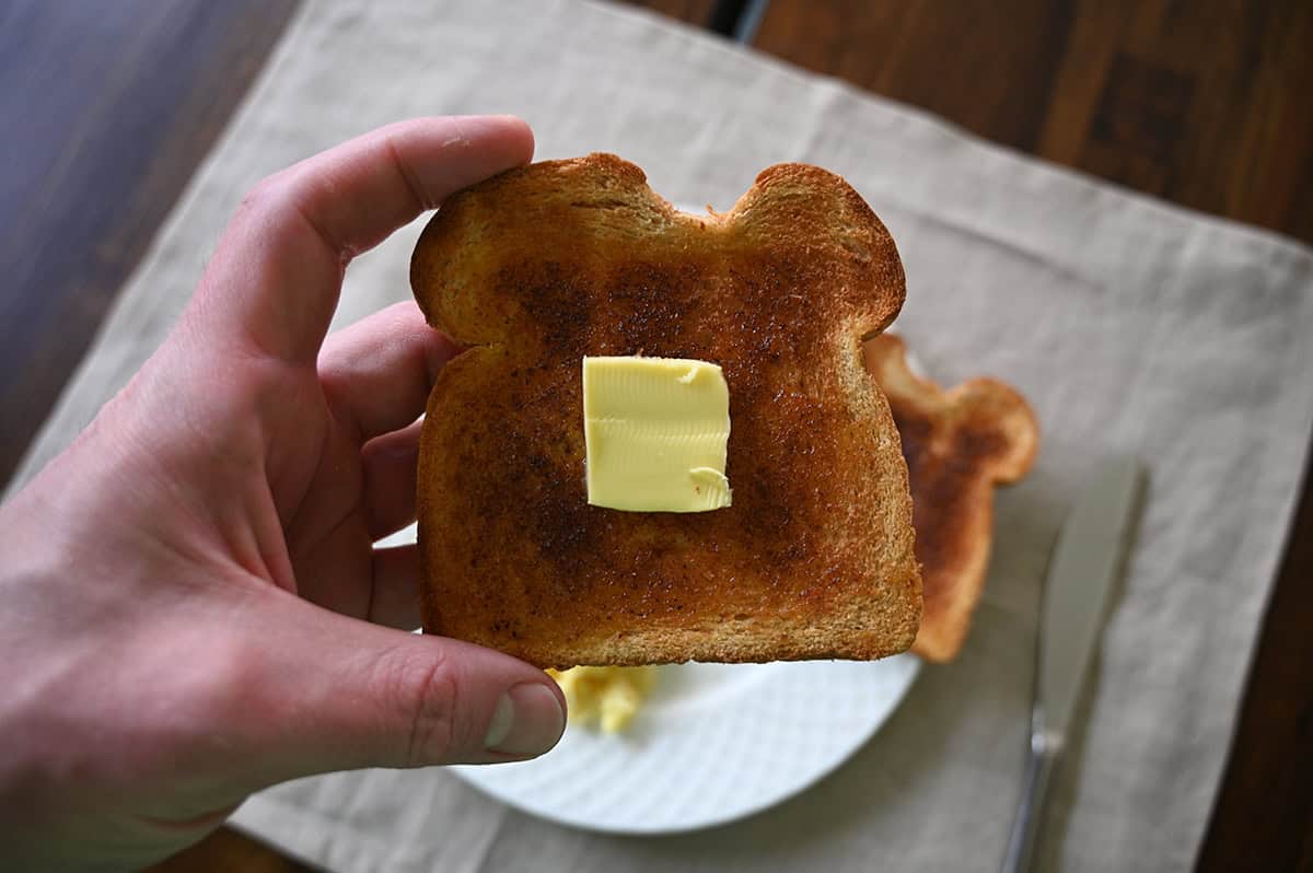 Closeup image of a hand holding a piece of toast and the toast has a large square of butter on it.