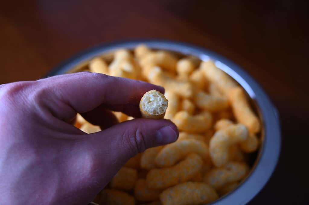 Sideview closeup image of a hand holding one puff with a bite taken out of it so you can see the middle close to the camera with a bowl of puffs in the background