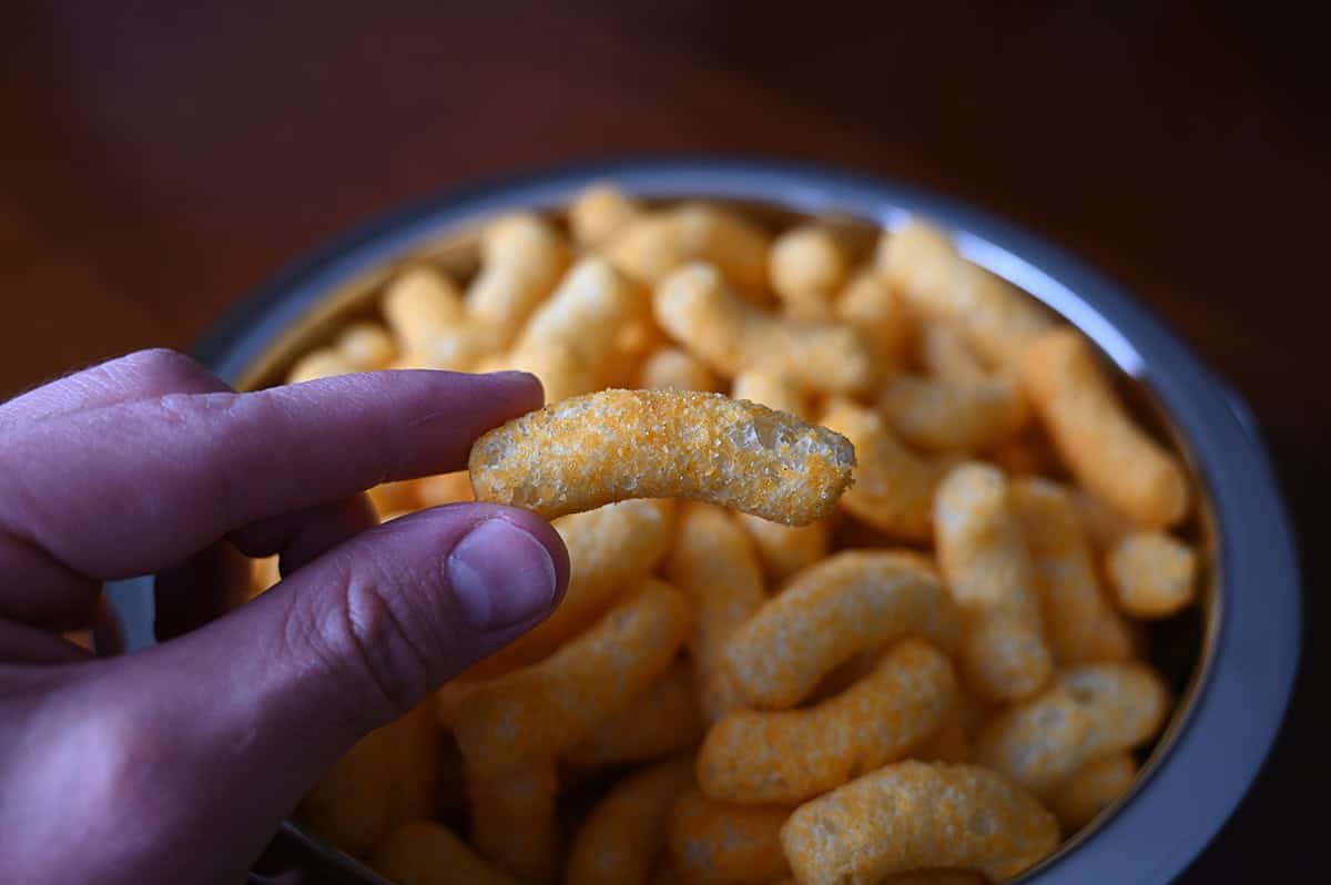 Sideview closeup image of a hand holding one puff close to the camera with a bowl of puffs in the background.