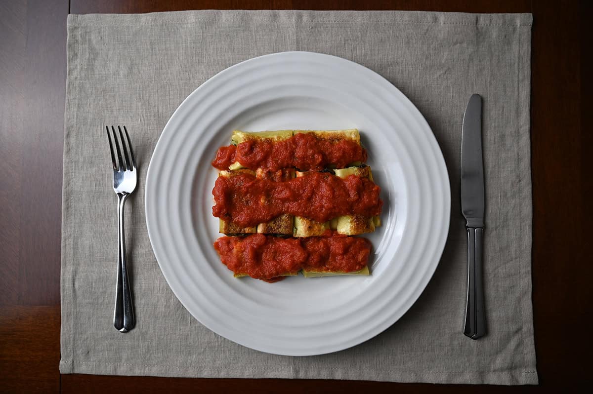 Top down image of a plate of cannelloni with marinara sauce on top and a fork and knife beside the plate.