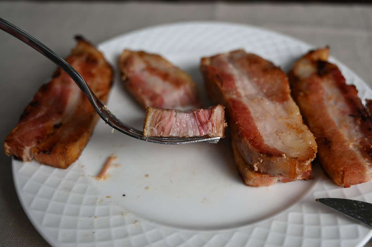 Closeup image of a fork with one bite of pork belly on the fork. In the background of the image is a plate of pork belly.