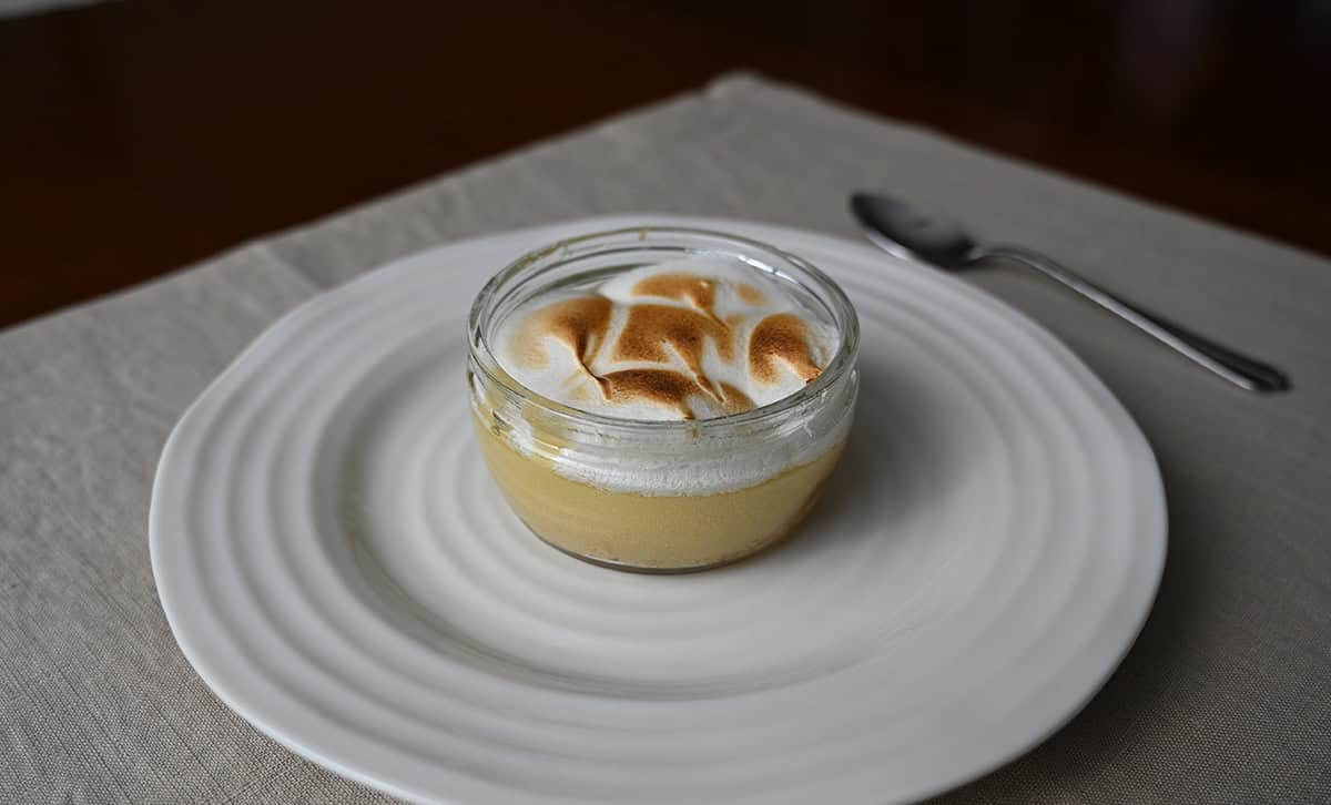 Sideview closeup image of one lemon meringue pie ramekin served on a white plate with a spoon beside it.