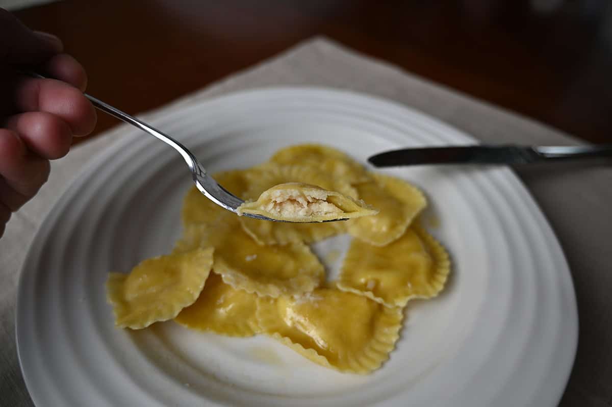 Closeup image one piece of ravioli cut in half resting on a fork so you can see the center with a plate or ravioli in the background. 