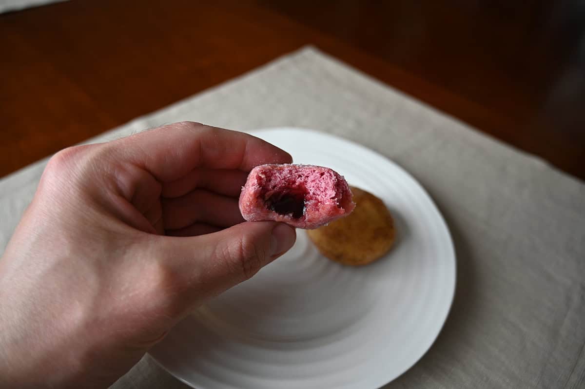 Image of a hand holding one red berry mini donut with a bite taken  out of it so you can see the middle. 