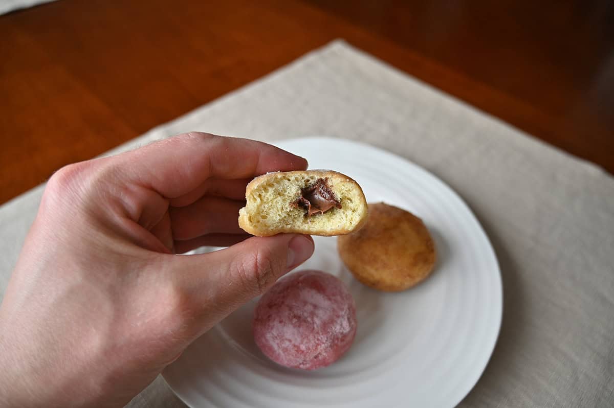 Image of a hand holding one chocolate hazelnut mini donut with a bite taken  out of it so you can see the middle. 