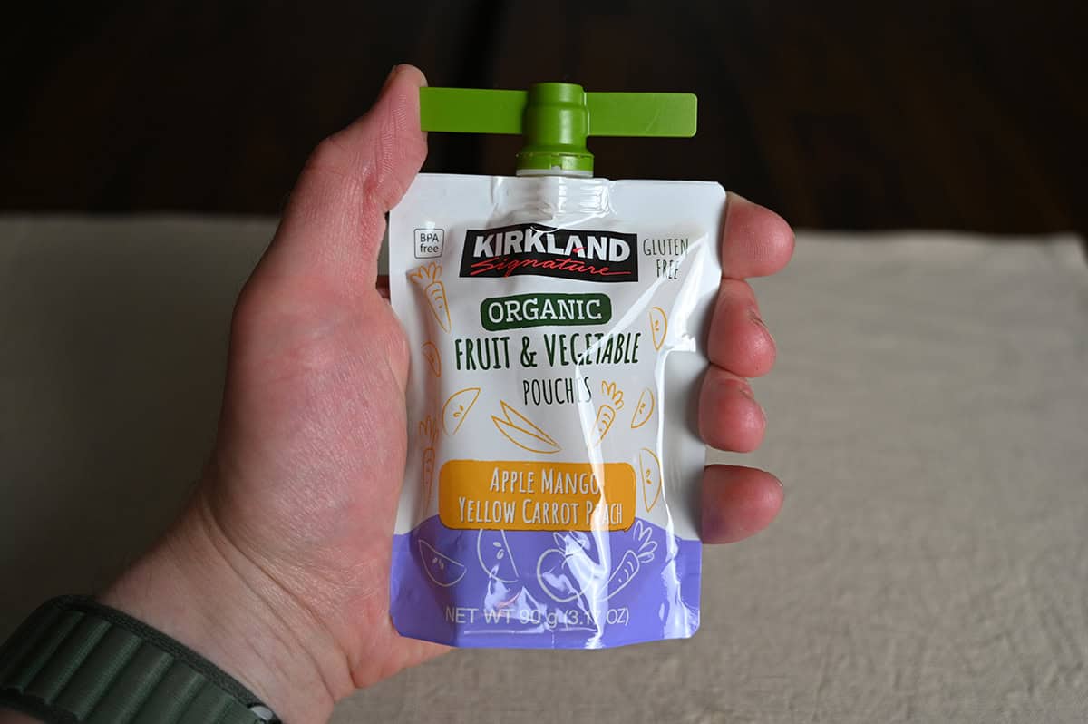 Image of a hand holding one Kirkland Signature Organic Apple Mango Yellow Carrot Pouch.