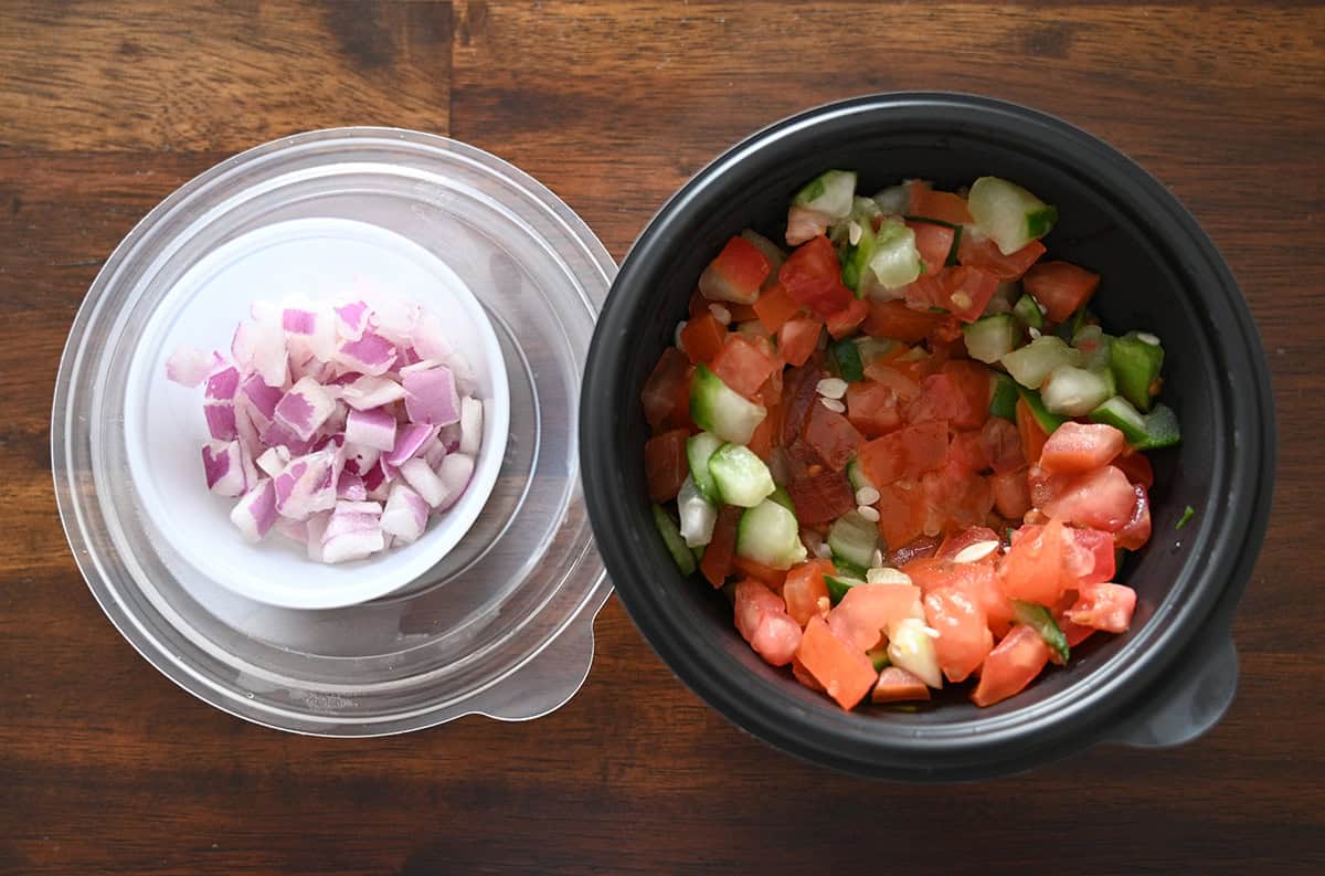 Top down image of an open container of cucumber and tomatoes and an open container of red onions sitting on a table. 