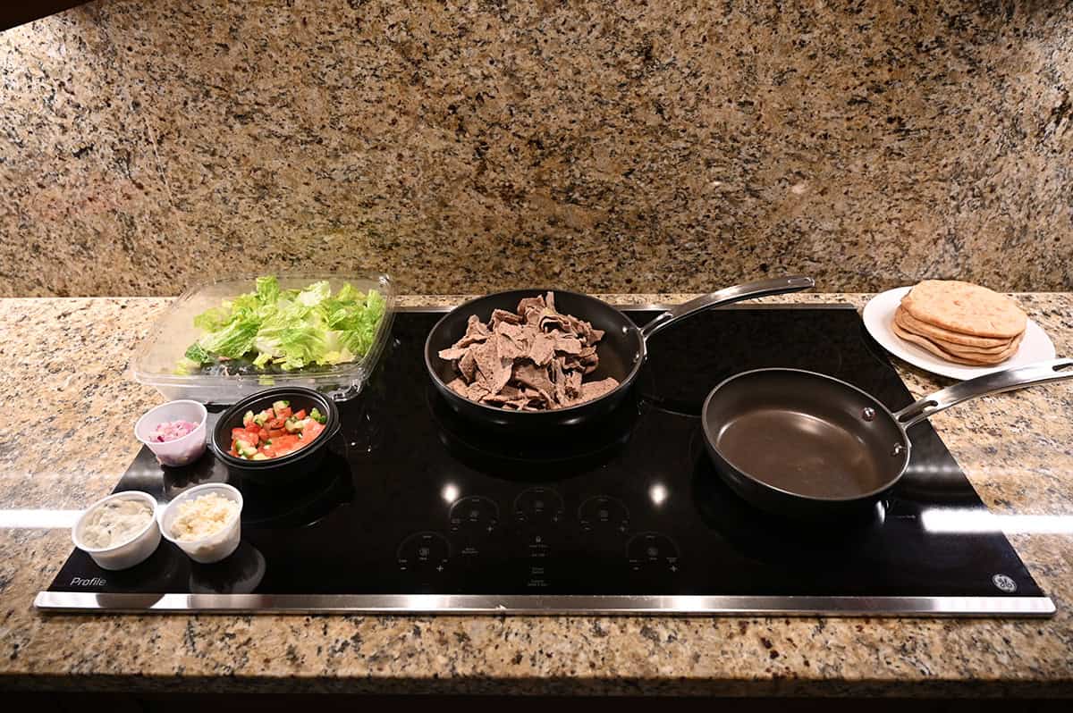 Image of a stovetop with all of the ingredients in the gyro kit surrounding the stove top, there is meat cooking in  a pan on the stovetop.