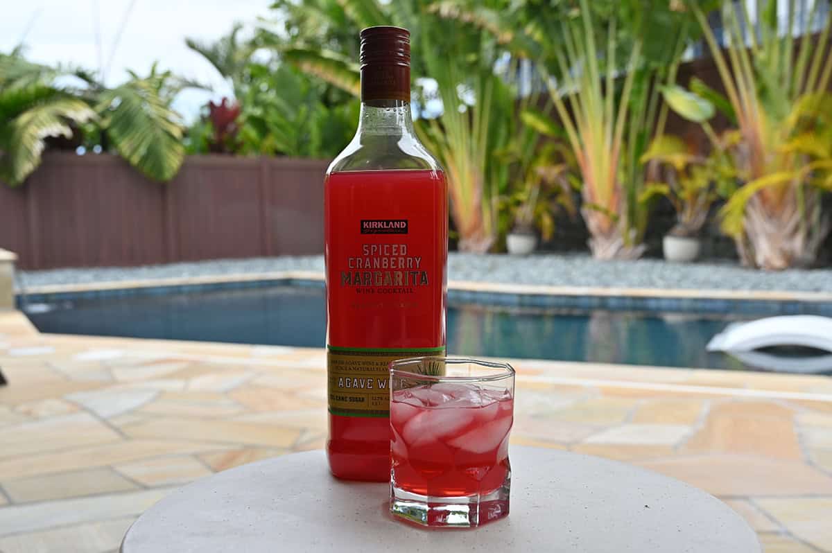 Image of the spiced cranberry margarita bottle standing on a table beside a glass of poured cocktail with a pool in the background.