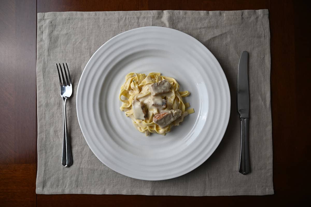 Top down image of a fettucine alfredo with chicken served on a white plate beside a fork and a knife.