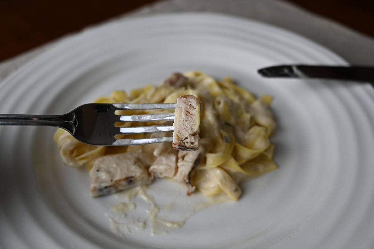 Closeup image of a fork with a piece of chicken on it, in the background is a plate of fettuccine.