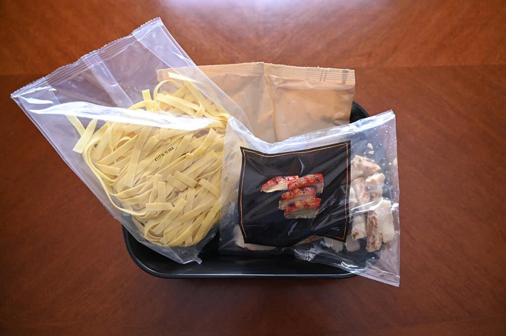 Top down image of everything that comes with the pasta. There is pasta, sauce and chicken in plastic bags sitting inside a tray.