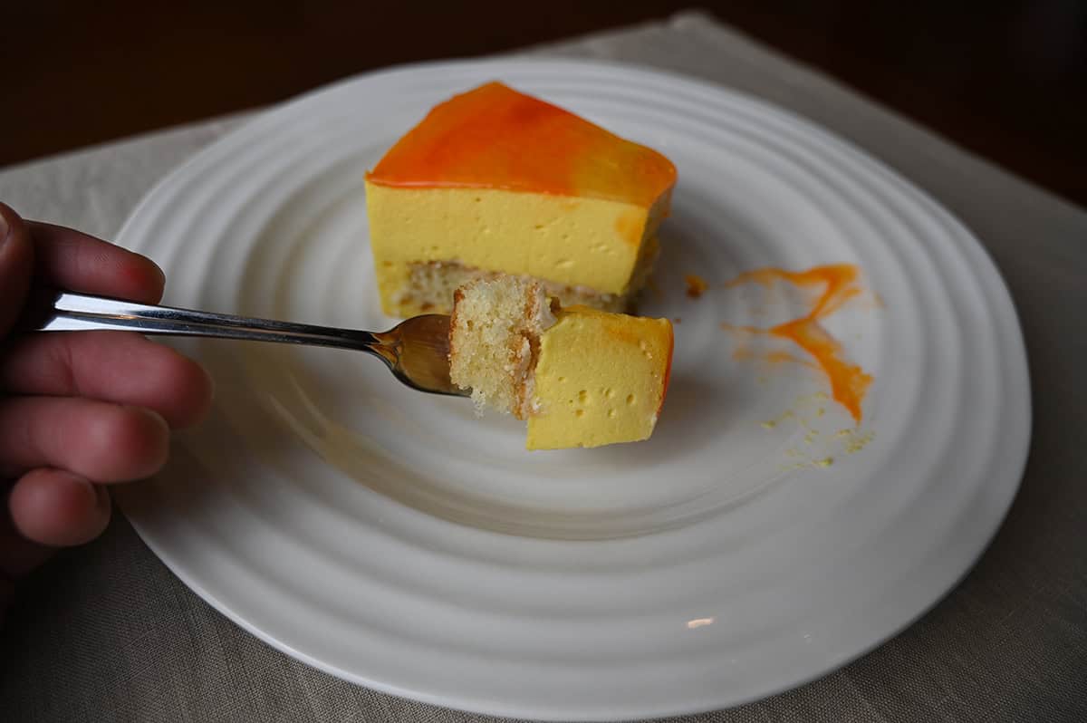 Closeup image of a fork with a bite of cake on it, in the background behind the fork is a slice of cake.