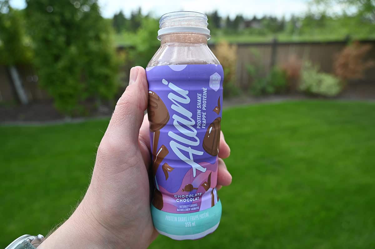 Closeup image of a hand holding one Alani protein shake close to the camera, in the background is trees and grass.