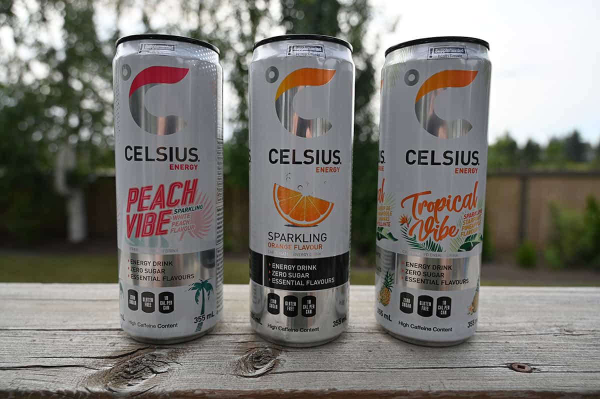 Image of three Celsius energy drinks sitting on a deck outside unopened in peach, orange and tropical flavor.