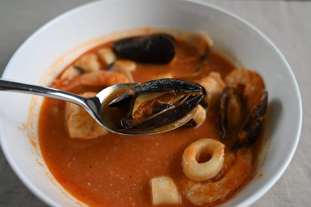 Closeup image of a spoon of cioppino hovering over the bowl of it. There is a clam on the spoon. 