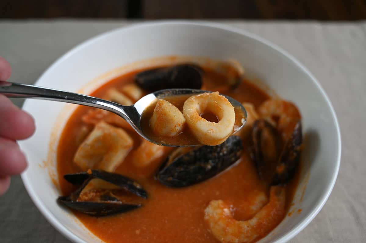 Closeup image of a spoon of cioppino hovering over the bowl of it. There is scallops and squid on the spoon. 