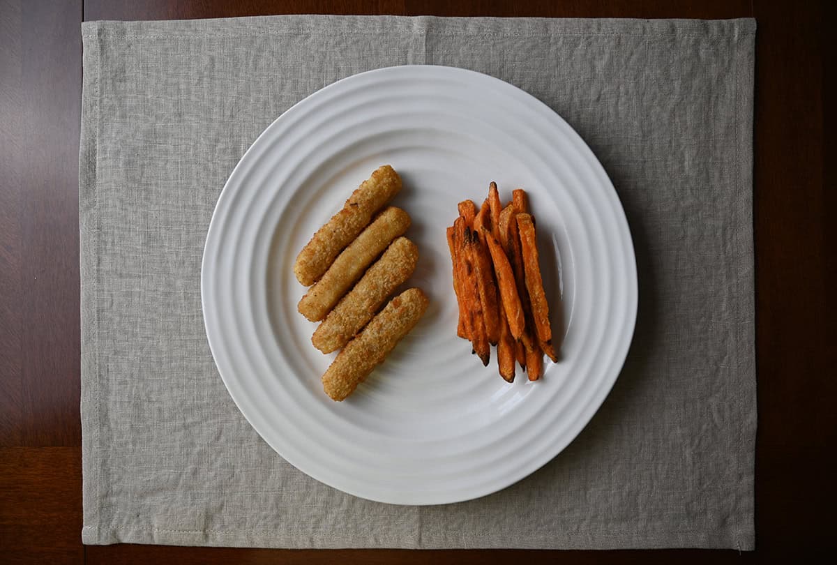 Top down image of four fish sticks cooked and served on a white plate beside some fries.