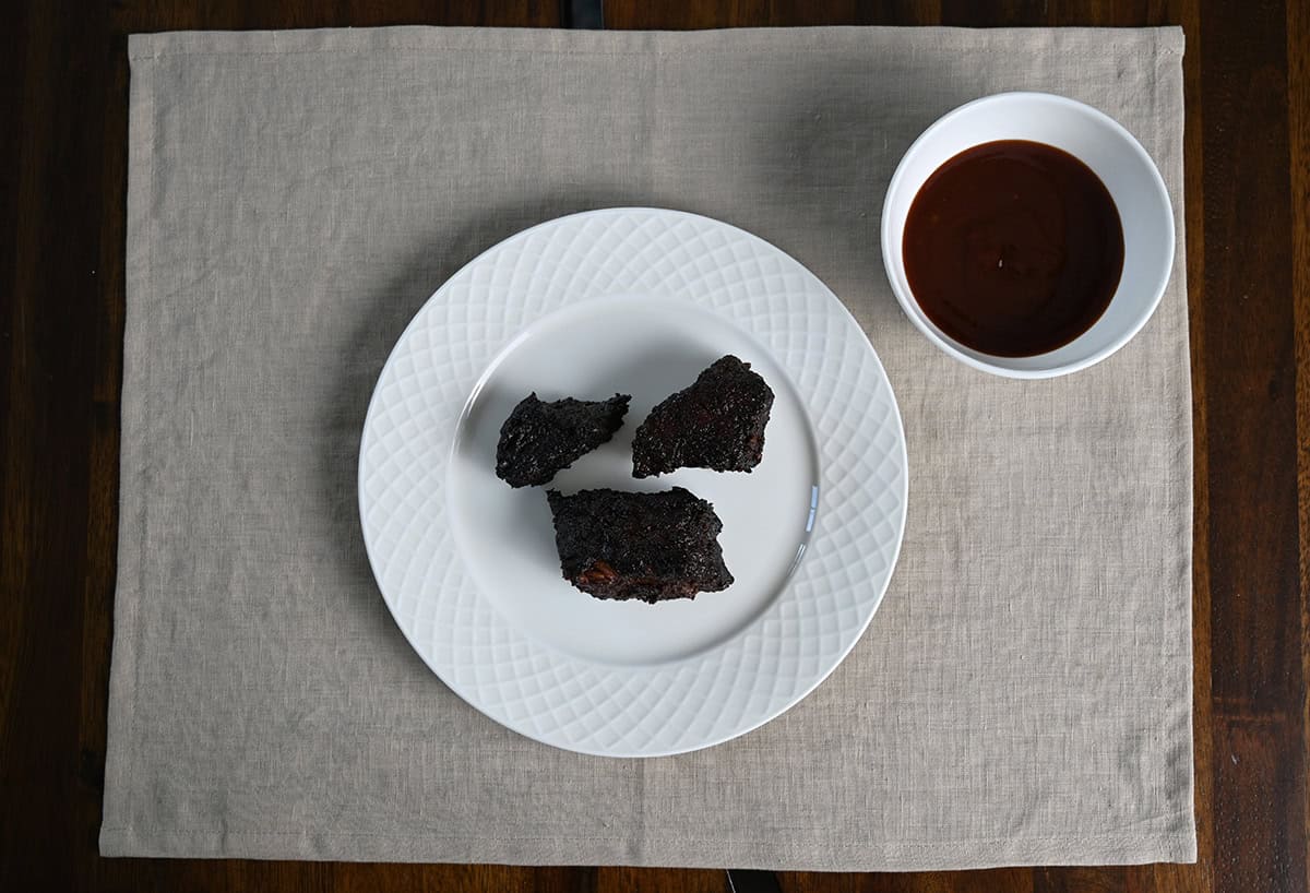 Top down image of three brisket burnt ends sitting on a plate beside a bowl of barbecue sauce.