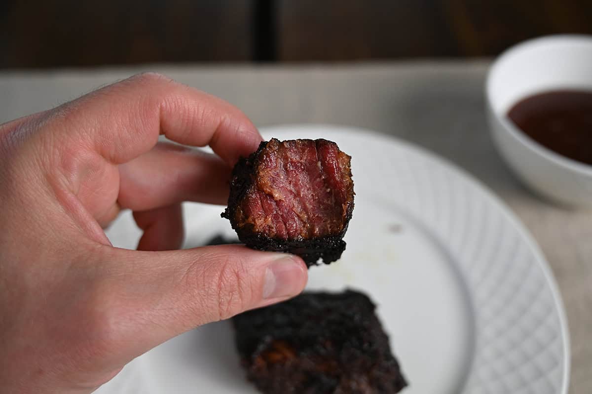 Image of a hand holding one small square brisket burnt end up close to the camera without barbecue sauce on it.