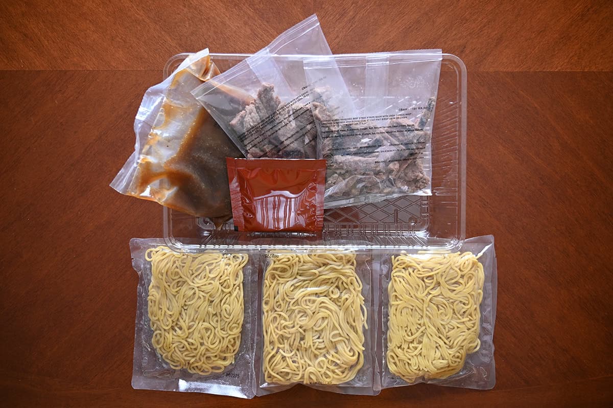 Top down image of everything that comes in the meal kit, three packs of noodles, sauce, veggie toppings and beef.