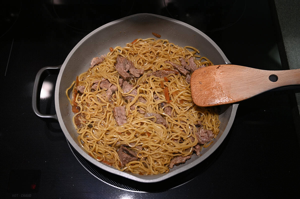 Top down image of a pan on a stovetop cooking a beef noodle stir fry and a spatula is stirring it.