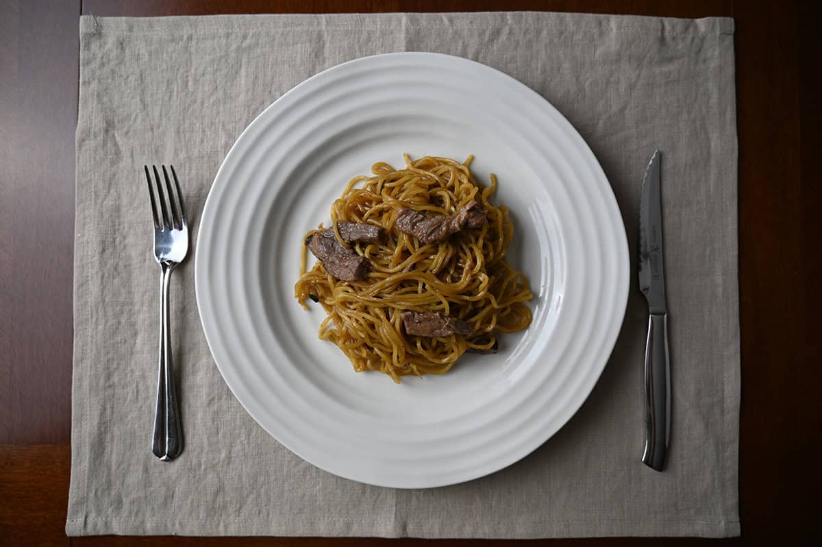 Top down image of a plate of beef noodle stir fry with a fork and knife resting beside the plate.