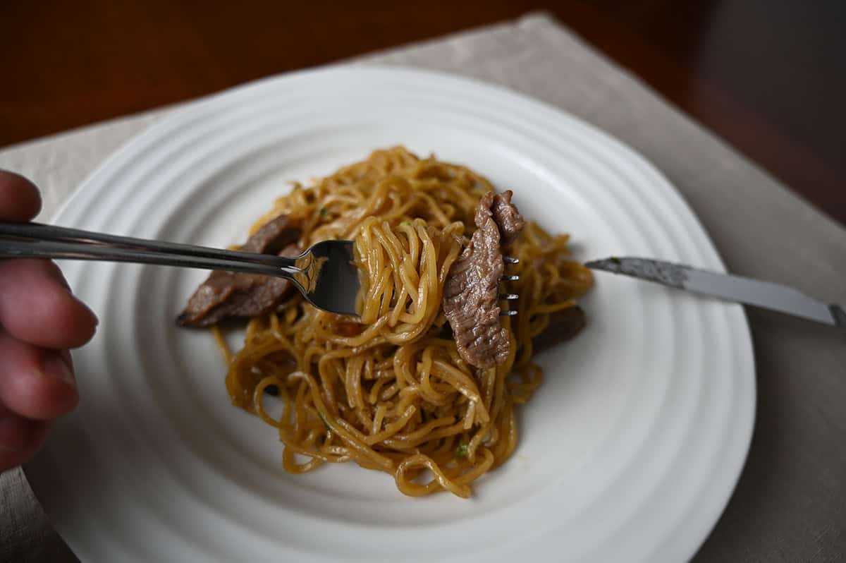 Sideview closeup image of a fork full of beef and noodles hovering over a plate of noodle stir fry.