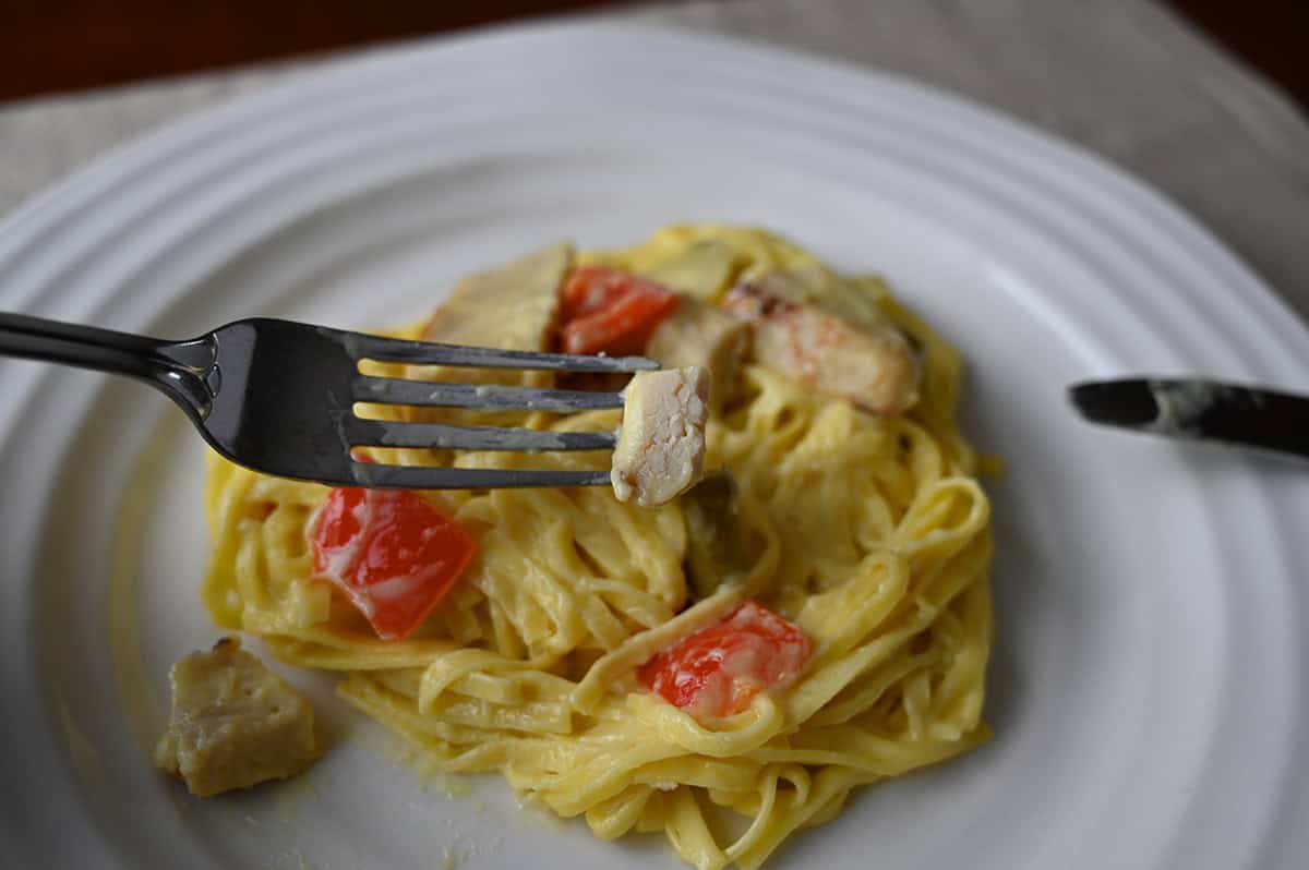 Closeup image of a fork with a piece of chicken on it hovering over a plate of pasta. 