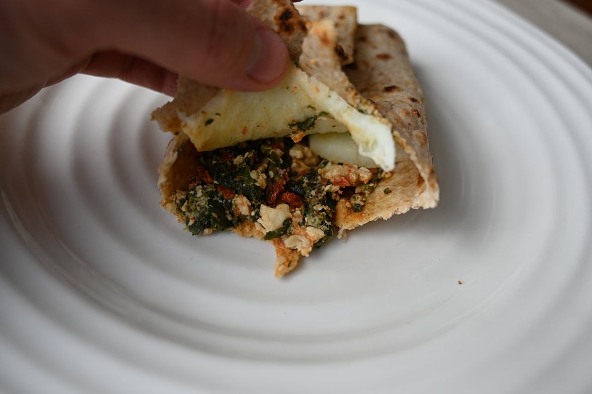 Image of a breakfast wrap served on a white plate with a hand holding one edge of the wrap so you can see the egg, spinach, sun-dried tomato and feta in the center.