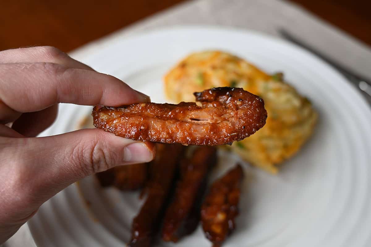 Closeup image of a hand holding one rib end close to the camera with a baked potato and more ribs ends on a plate in the background.