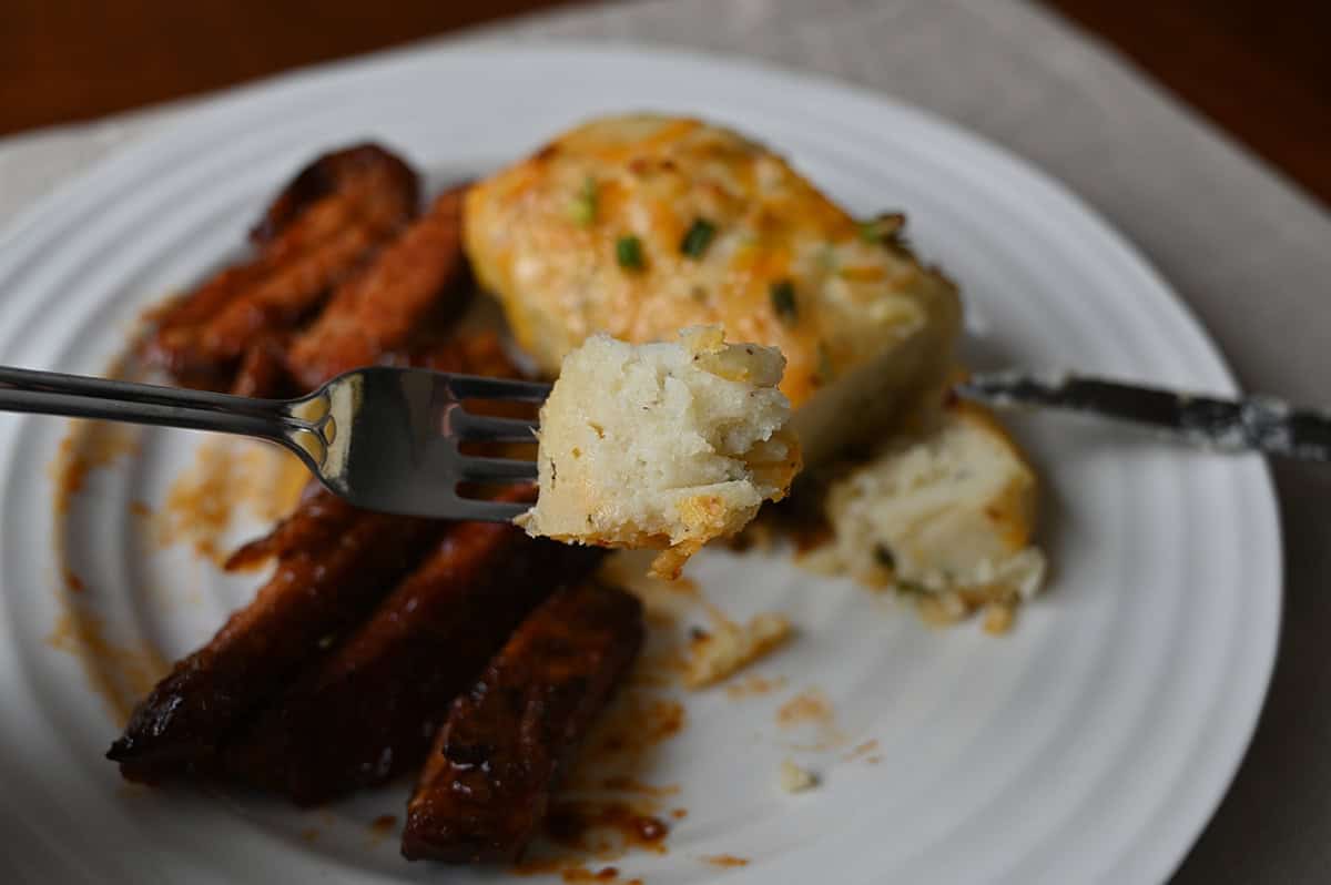 Closeup image of a fork close to the camera with a bite of baked potato on it. In the background of the image is rib ends and baked potato.