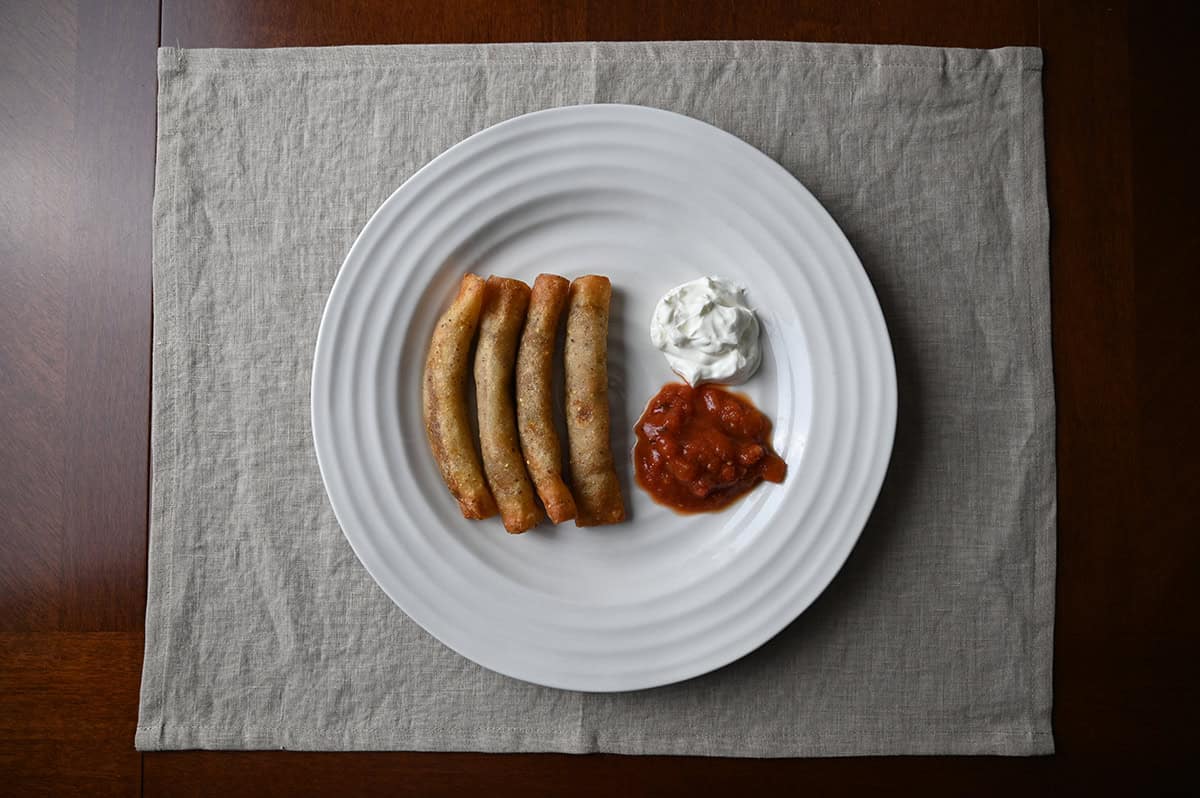 Top down image of four rolled tacos served on a white plate beside sour cream and salsa.