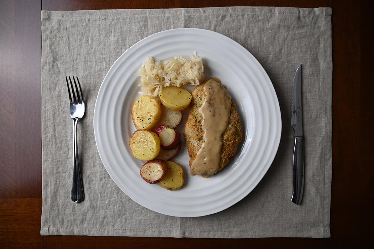 Image of a plate with pork schnitzel with gravy on top beside a side of potatoes and sauerkraut.