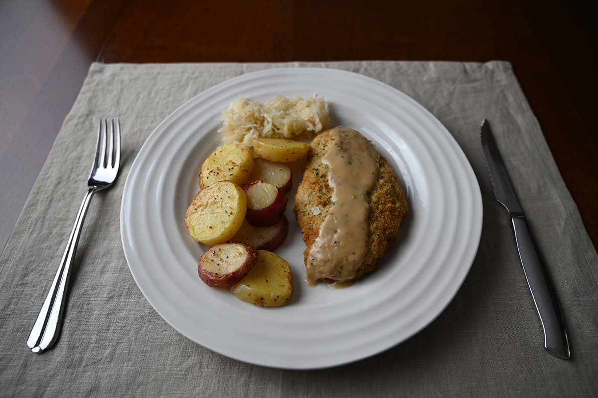 Image of a plate with pork schnitzel with gravy on top beside a side of potatoes and sauerkraut. The plate has a fork and knife beside it.