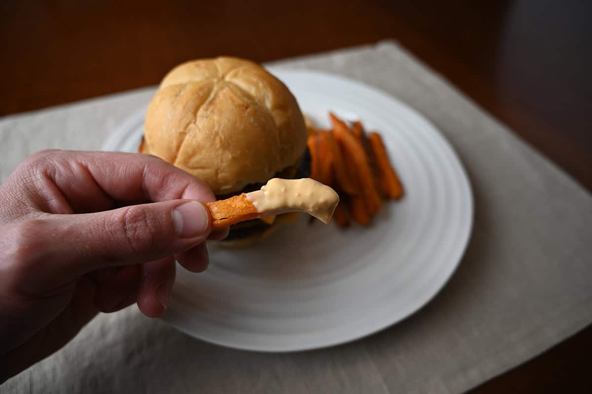 Closeup image of a hand holding a fry dipped in aioli with a plate of fries and a burger in the background.