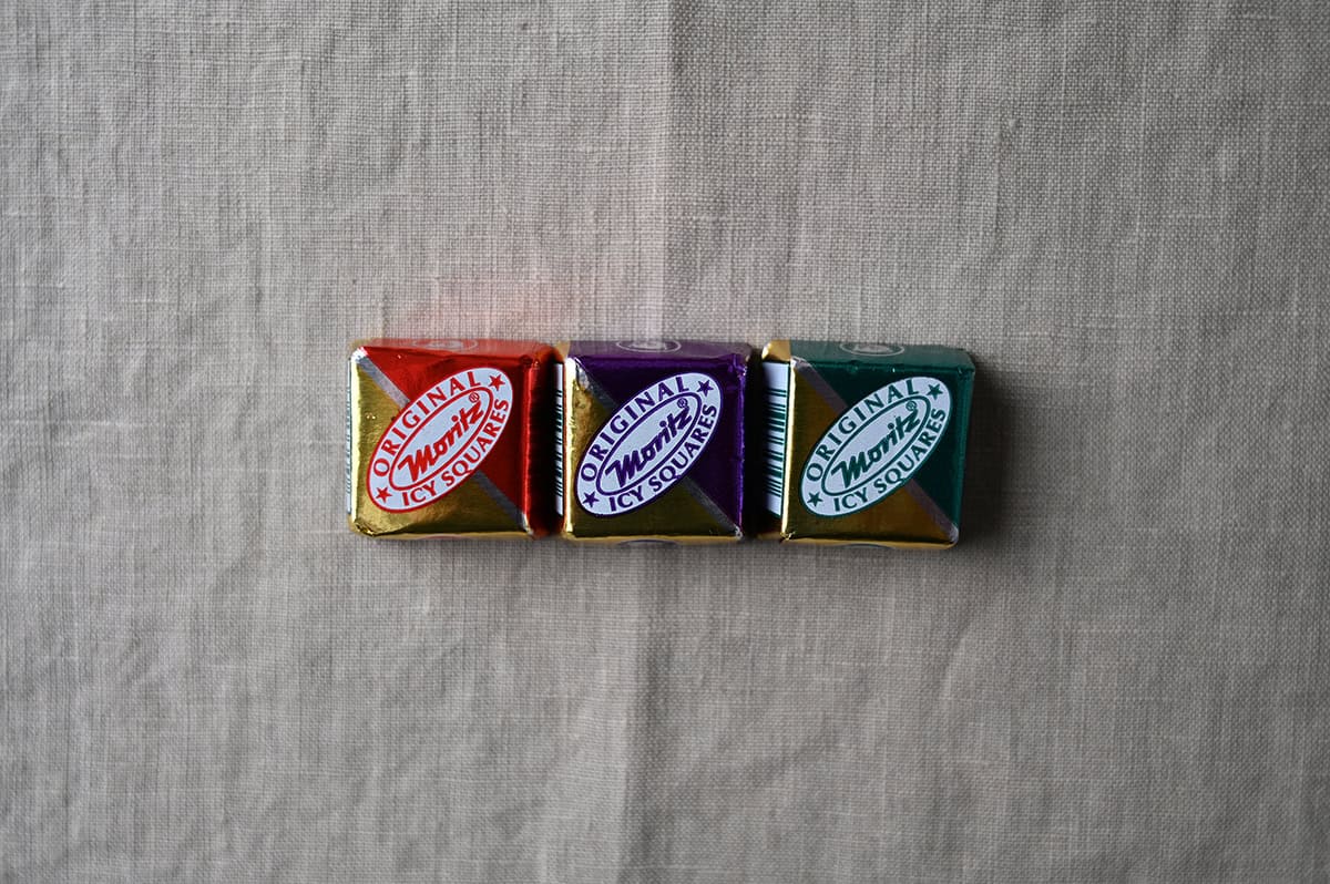 Image of three wrapped icy squares sitting on a table, there is a red one, a purple one and a teal one.