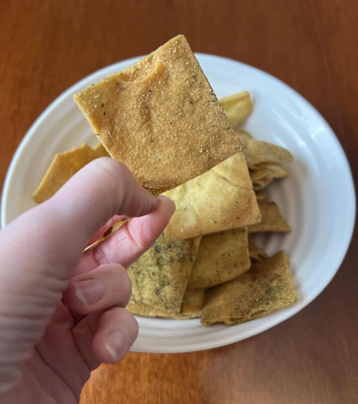 Image of a hand holding one pita chip close to the camera with a bowl of pita chips in the background.