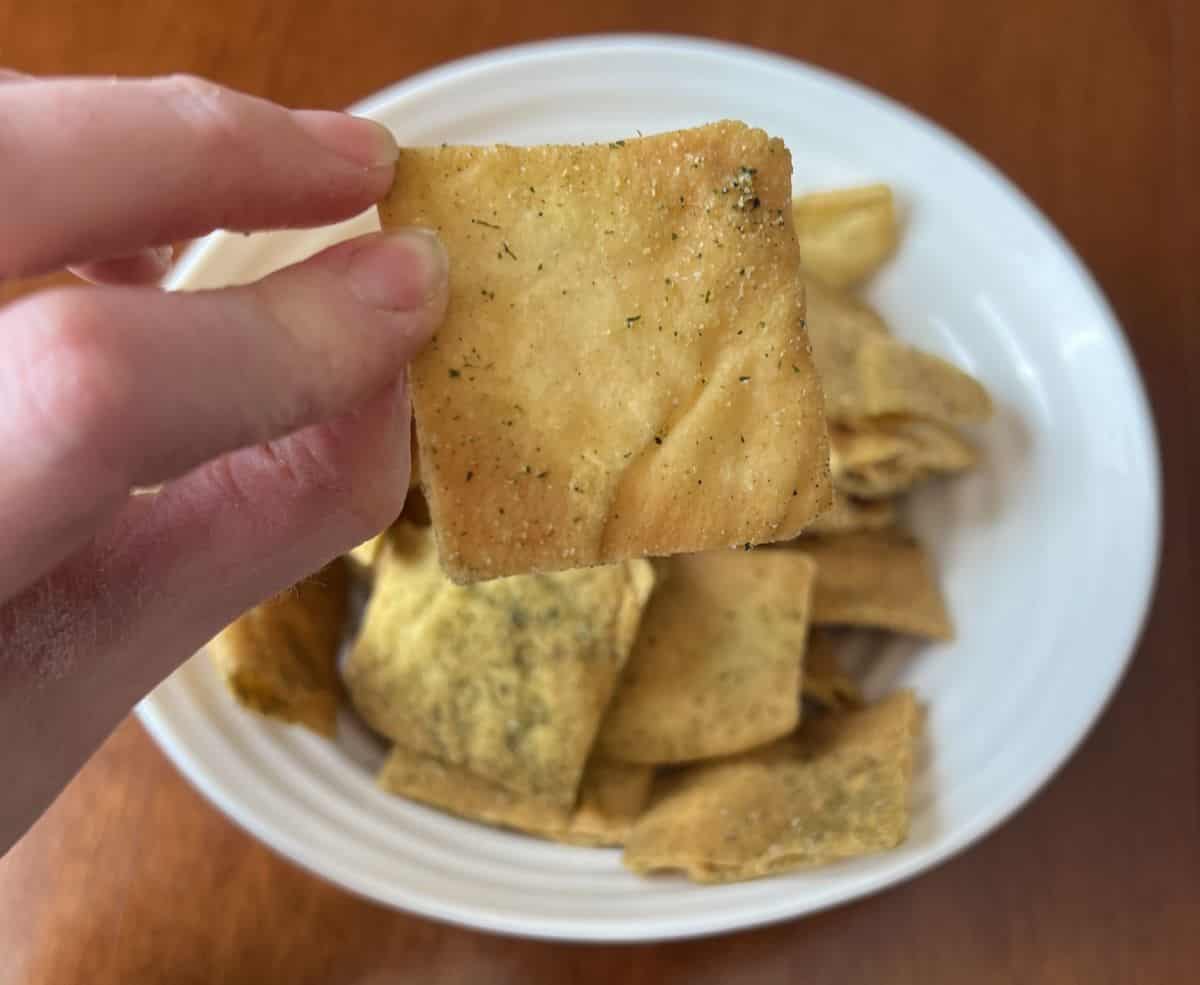 Image of a hand holding one pita chip close to the camera with a bowl of pita chips in the background.