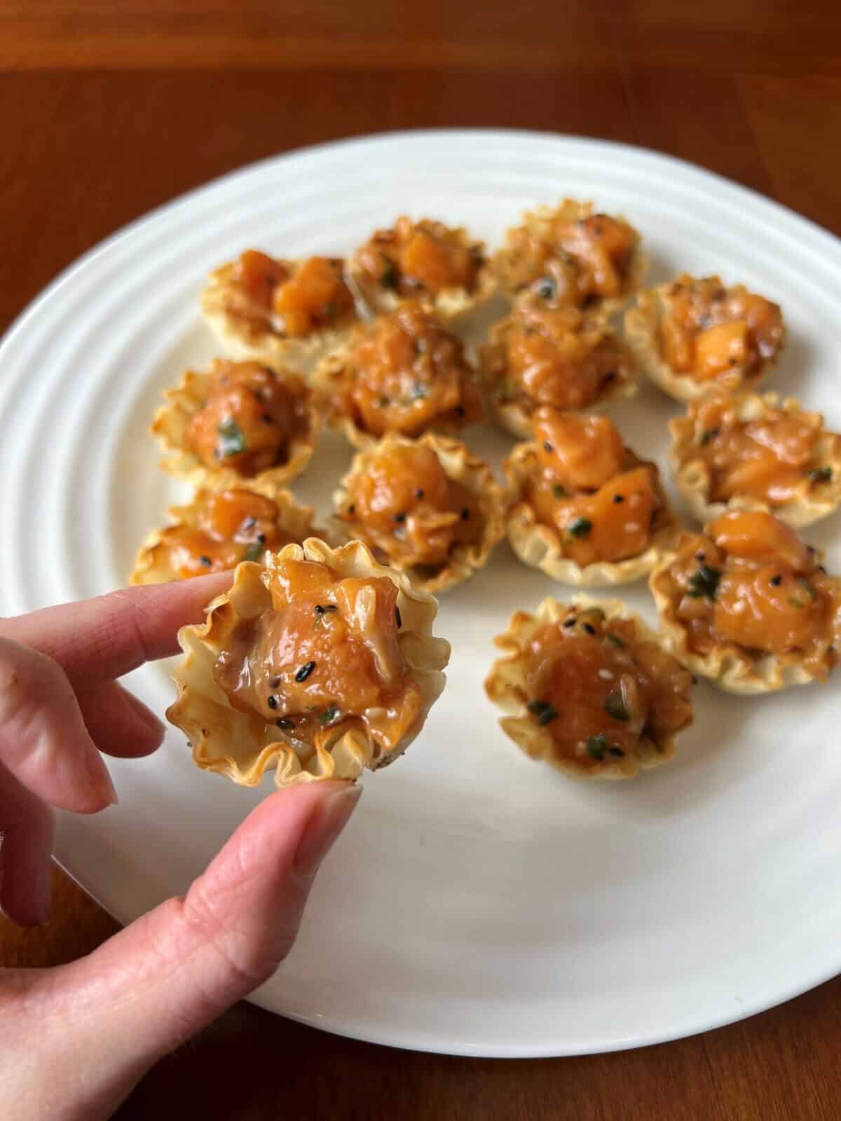 Image of a hand holding one salmon poke bite close to the camera with a plate of salmon poke bites in the background. 