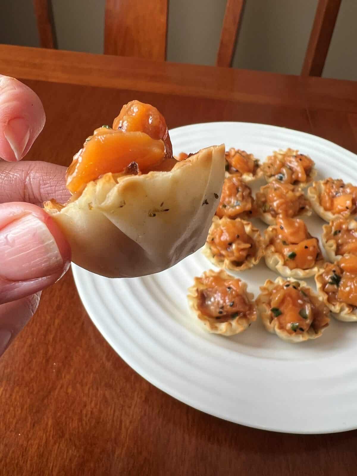 Closeup image of a hand holding one salmon poke bite shell so you can see the depth of the phyllo pastry shell. 