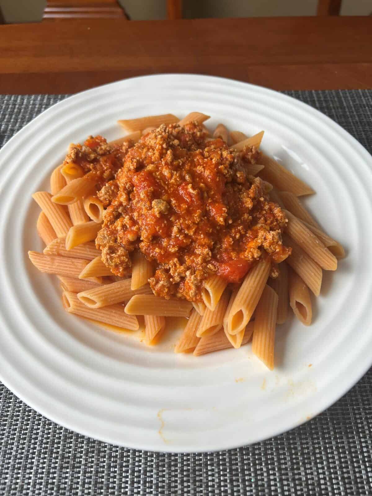 Top down closeup image of a plate of red lentil penne with Bolognese sauce on top.