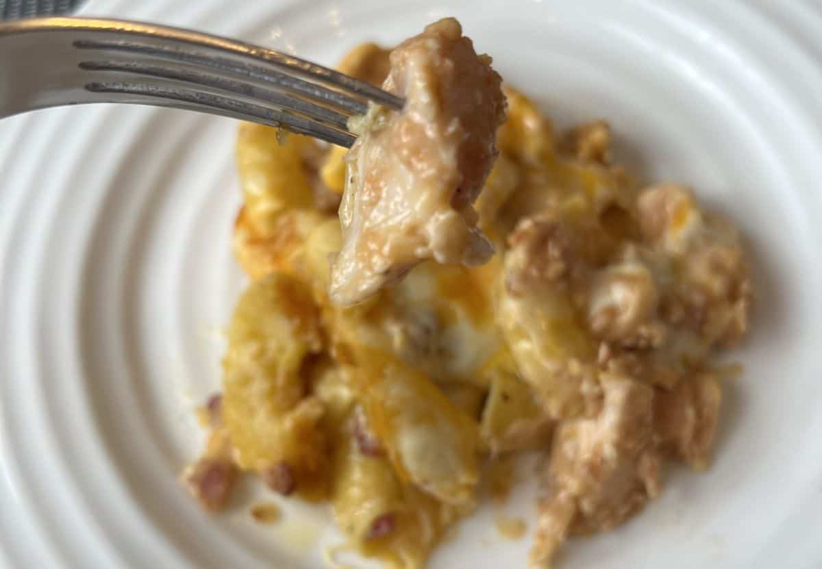 Image of a fork with a bite of chicken on it hovering over a plate of chicken and bacon mac and cheese.