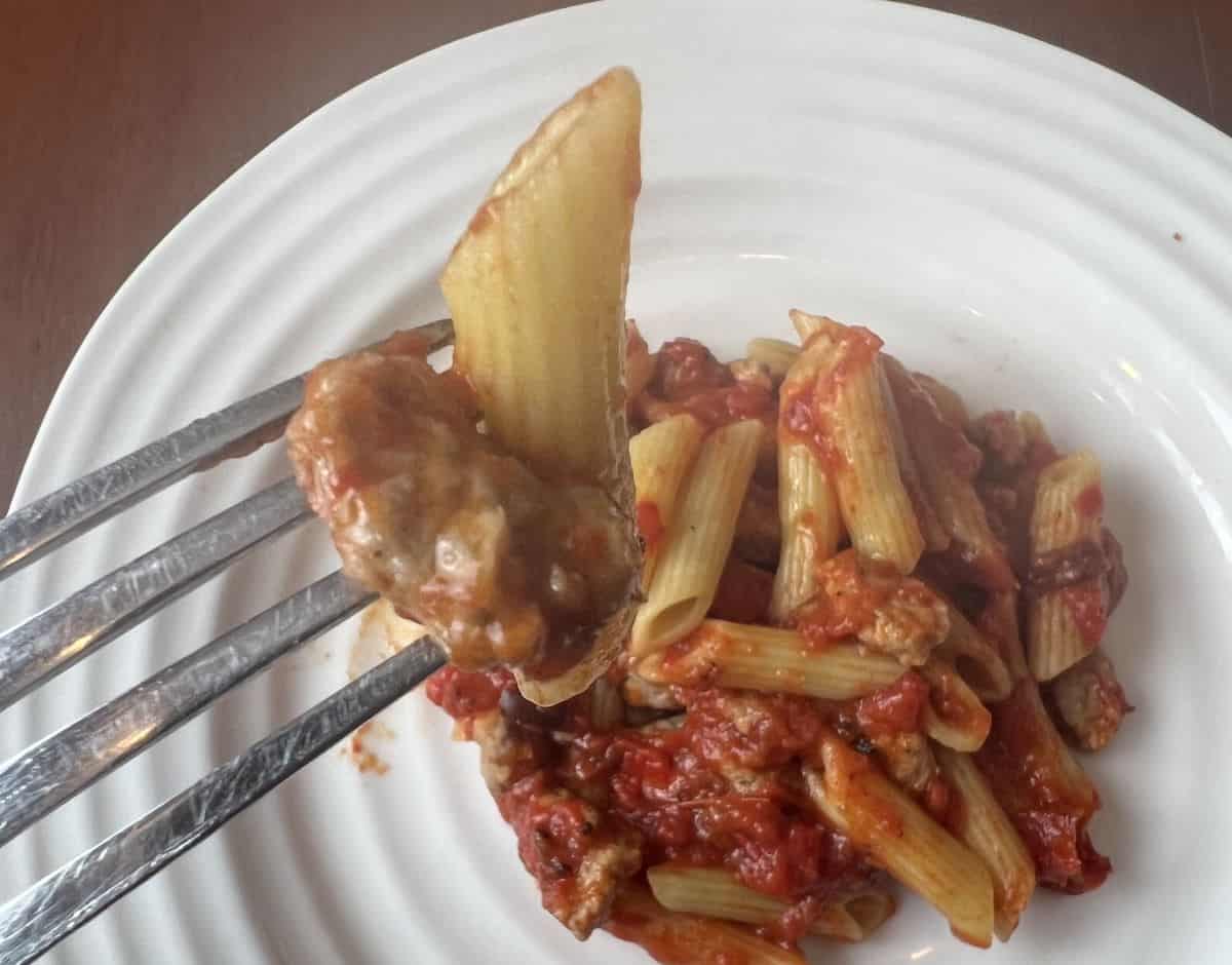 Closeup image of a fork with a penne noodle on it and a piece of Italian sausage. In the background is a plate of pasta. 